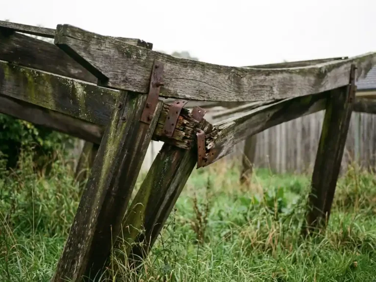 Verweerde houten pergola met hangende balk en losse bevestigingen in achtertuin met overgroeid gras